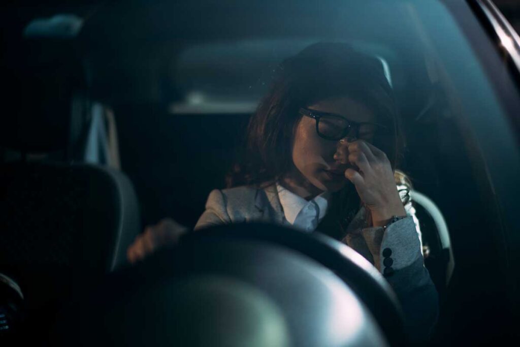 a woman pinches the bridge of her nose while behind the wheel of a car after fall back daylight savings