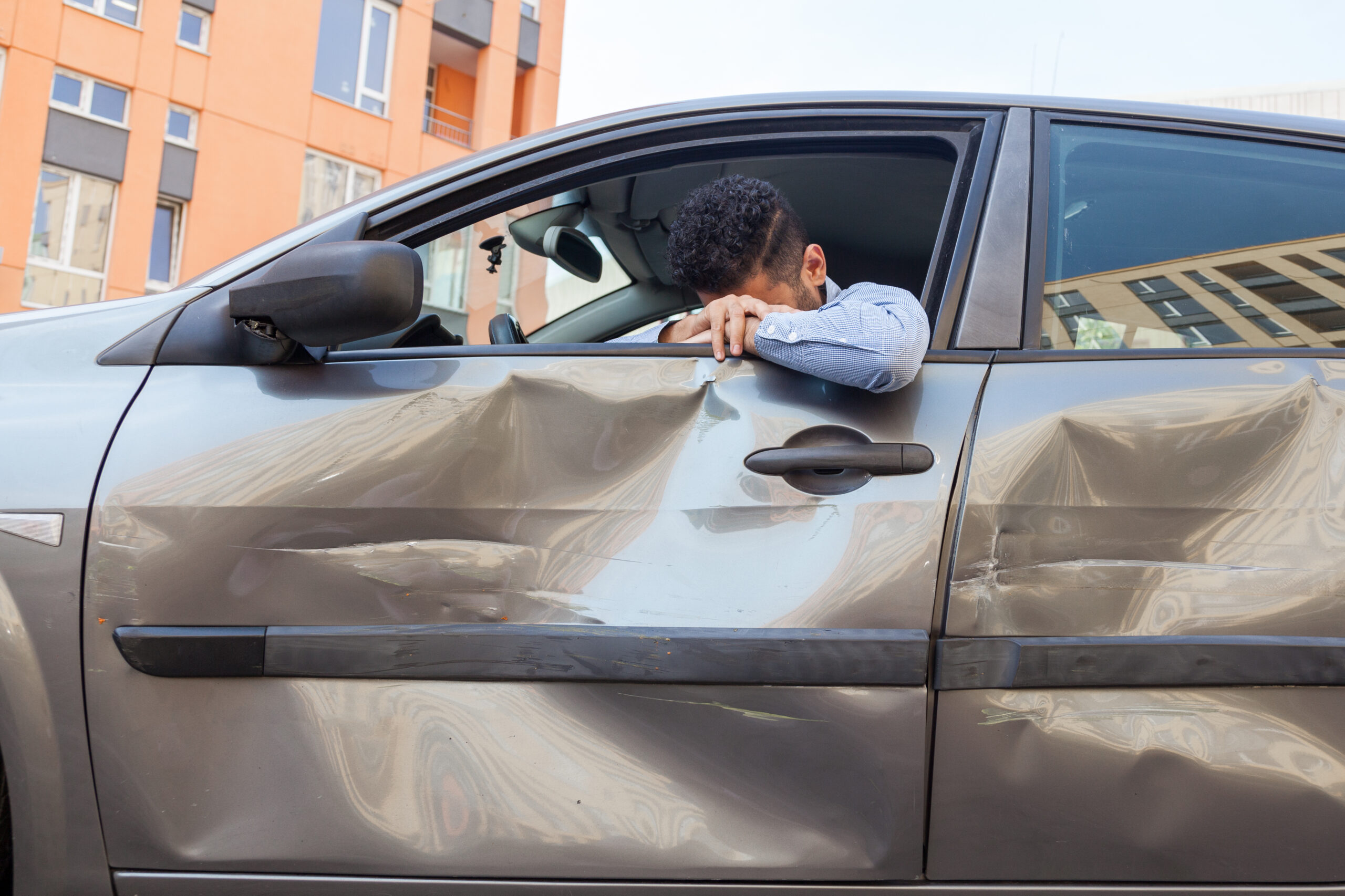 Sad man sitting behind the wheel of wrecked car and puts his head down, sobbing, scratches and dents on auto door.