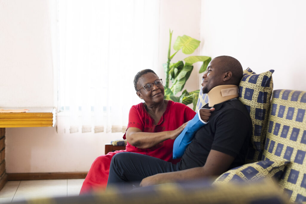 Senior woman helps adult son with injury to shoulder and neck sitting in living room after catastrophic injury accident