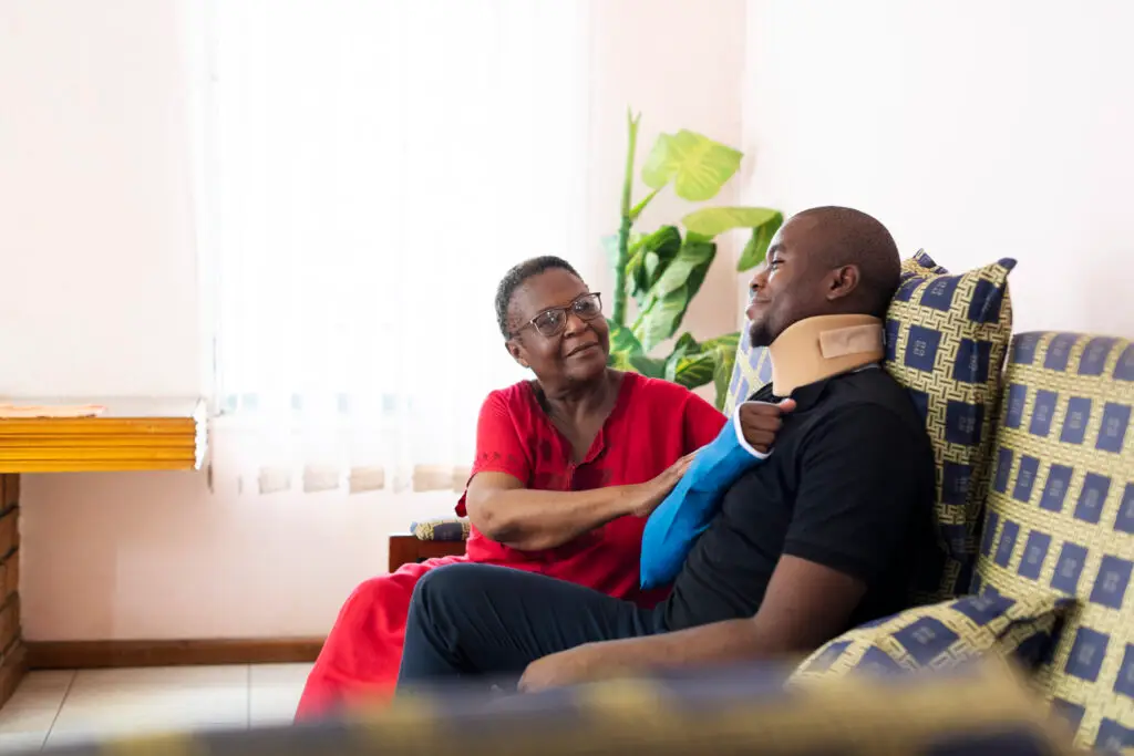 Senior woman helps adult son with injury to shoulder and neck sitting in living room after catastrophic injury accident